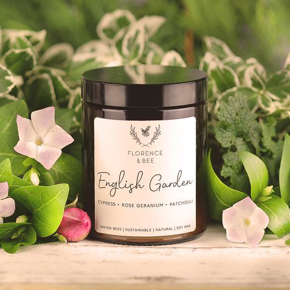 A dark amber glass candle jar with a black lid and a white label reading 'FLORENCE & BEE English Garden', styled on a wooden surface with green leaves and pale pink flowers.